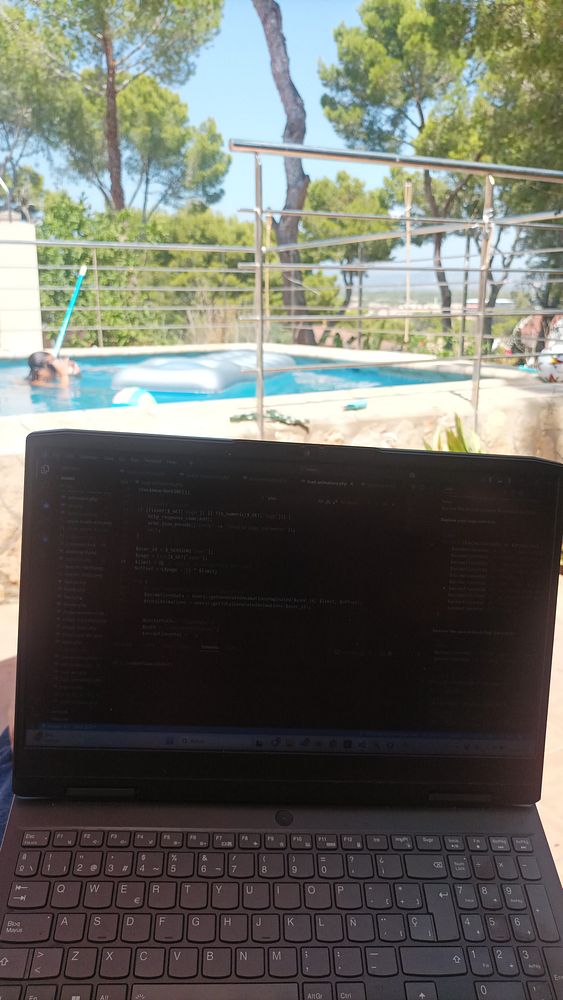 A laptop is positioned outdoors with a view of a swimming pool and trees in the background.
