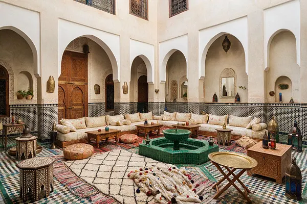 A Moroccan-style backyard courtyard with white stucco walls, pointed arches, patterned tile wainscoting, and a central green tiled fountain. Low cream cushions line the perimeter, surrounded by carved wooden tables, lanterns, poufs, and layered rugs in warm earth tones and jewel accents.