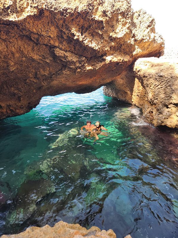 Two people snorkeling together in clear water beneath a large rocky overhang.