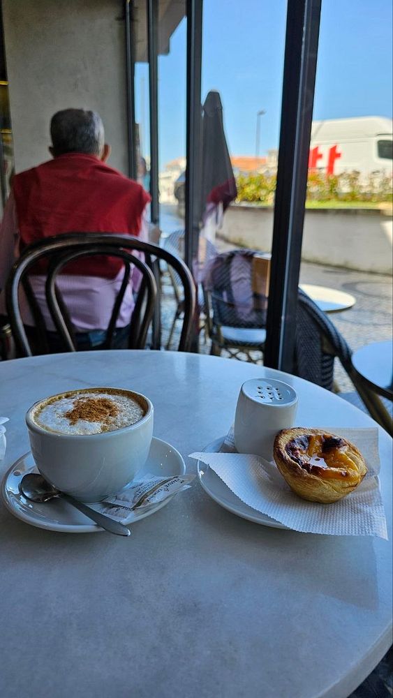 A pastel de nata with a crispy, golden-brown crust and creamy custard filling in a white paper cup, placed on a speckled black and beige ceramic plate.