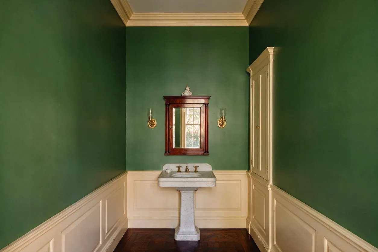 A traditional powder room with deep green walls above cream wainscoting, a dark wood framed mirror, and a white marble pedestal sink centered on the back wall. Brass wall sconces flank the mirror, while a tall built-in cabinet in a soft cream finish adds storage on the right.