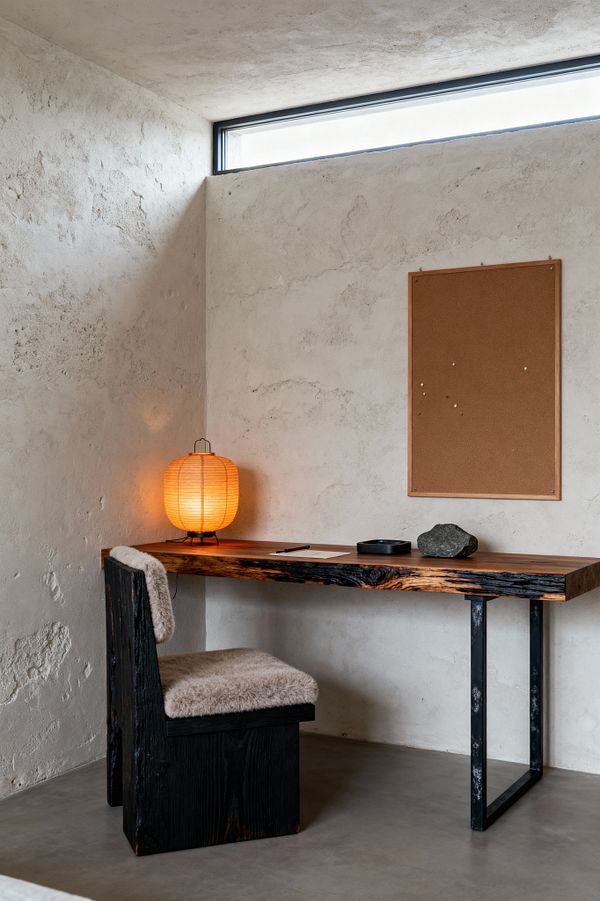 A minimalist home office corner features a charred wood desk with a blackened steel leg set against textured off-white lime plaster walls. A narrow clerestory window brings daylight, while a glowing paper lantern warms the scene. A chunky chair in matching charred wood has beige shearling cushions. On the desk are a sheet of paper with a pencil, a black dish, and a single stone beneath a simple cork pinboard.