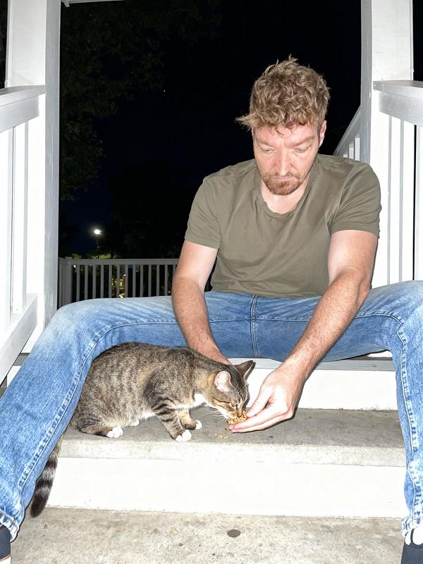 A man sits on a porch step feeding a cat at night.