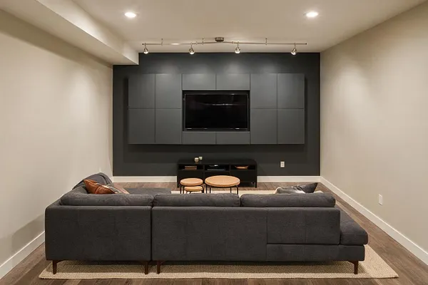A modern basement media room with a dark gray sectional sofa on a neutral rug facing a wall-mounted TV. The back wall features matte charcoal built-in cabinets, a black media console, and track lighting, while light walls and warm wood-look flooring balance the room.