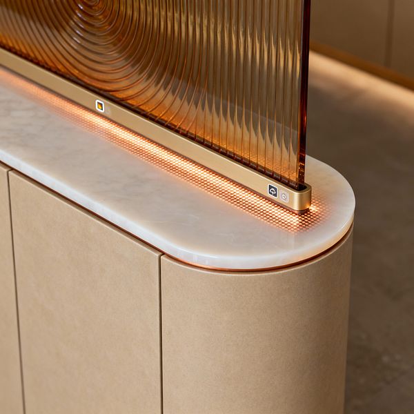 Close-up of a futuristic kitchen island corner with seamless sand‑beige cabinetry and a polished white marble top with a rounded bullnose edge. A ribbed, amber‑tinted glass screen with concentric grooves sits in a champagne‑gold frame showing small touch icons, above a perforated copper grille that emits a warm LED glow along the stone edge. The design is handleless and smooth, emphasizing clean lines and gentle curves.