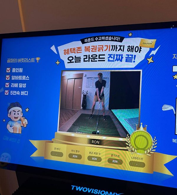 An indoor golf simulator setup in a dark room with a projected golf course on a large screen and golf clubs along the side.