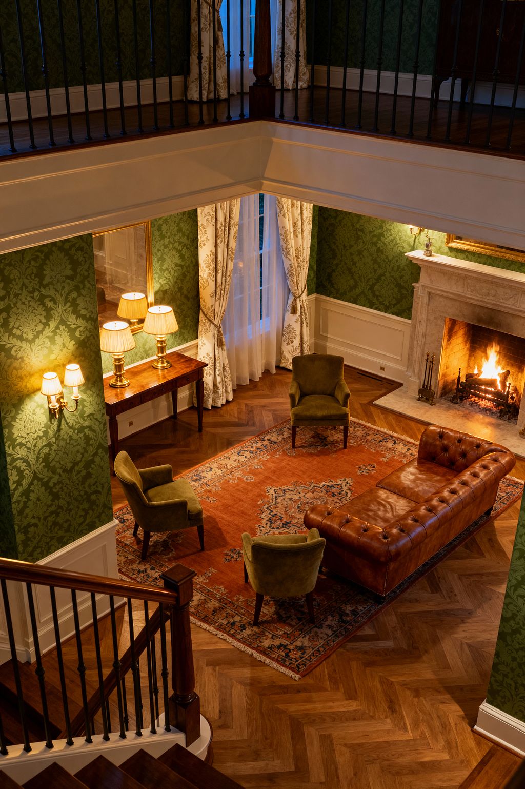 Overhead view of a double-height traditional living room with green damask wallpaper above white wainscoting. A carved marble fireplace burns brightly, facing a cognac tufted leather Chesterfield and three olive velvet armchairs arranged on a rust Persian rug over oak herringbone flooring. Floor-length cream patterned drapes frame tall windows, while brass sconces and pleated-shade table lamps on a wood console add warm light. A dark balustrade lines the mezzanine and staircase.