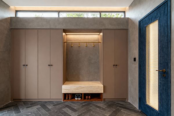 Contemporary mudroom with symmetrical taupe cabinets flanking a central niche. A thick travertine bench sits above walnut cubbies for shoes, with a brass rod and five hooks beneath a concealed LED strip. Walls are warm gray microcement and the floor is charcoal herringbone tile. A long clerestory window and cove lighting add daylight, while a deep blue door with ribbed glass and a brass lever punctuates the right side.