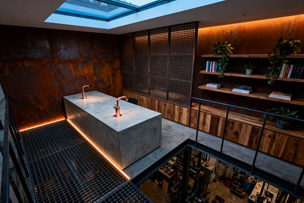 An upper-level industrial home office features a large concrete desk with two copper task lamps set on a steel grating floor edged with warm LED lighting. Rusted corten-steel wall panels, perforated metal screens, and open hardwood shelves with books and potted plants surround the space. A wide skylight brings in daylight, black steel railings line the mezzanine, and the office overlooks a workshop below.