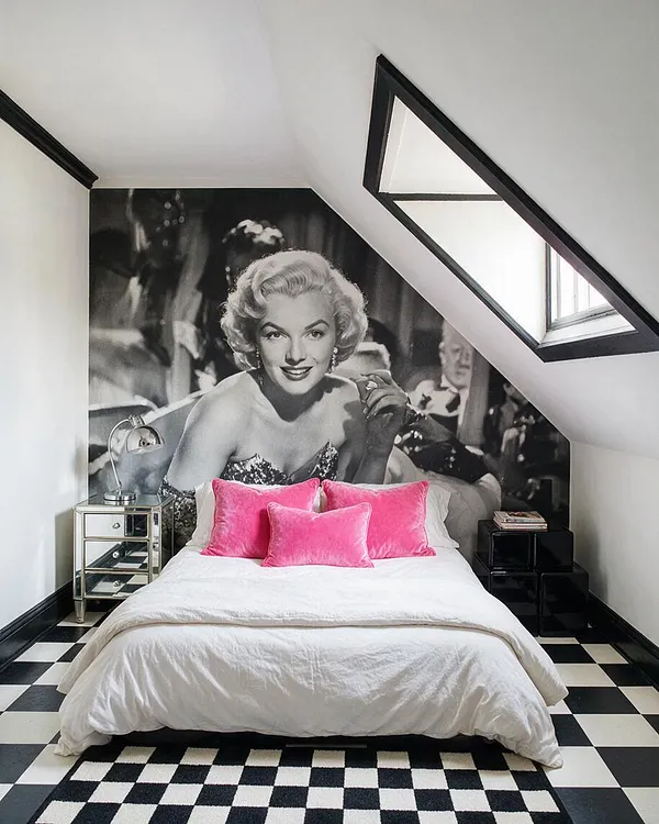 A small guest bedroom with a sloped ceiling features a large black-and-white portrait mural of Marilyn Monroe behind a low bed dressed in white bedding and bright pink velvet pillows. The room has a black-and-white checkered floor, mirrored bedside table, black stacked nightstands, and a small angled window with black trim.