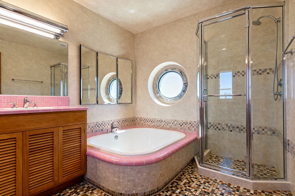 A mid-century bathroom features a white soaking tub wrapped with pink ceramic bullnose tile and a mosaic-clad apron beside a chrome-framed glass shower lined in beige tile with a small mosaic border. A warm wood louvered vanity with a pink mosaic countertop sits under tri-panel mirrored cabinets and a chrome fluorescent light, while a round porthole window above the tub brings daylight to a floor of multitone beige and brown mosaic tile.