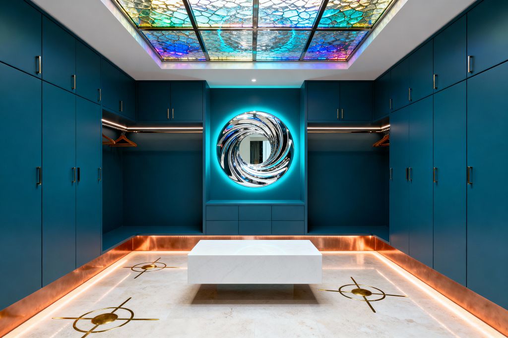Symmetrical mudroom lined with matte teal cabinets and brass handles, featuring open hanging bays with warm LED-lit rails and a copper base plinth with perimeter lighting. A white marble block bench rests on pale stone flooring inset with brass compass motifs, while a round chrome, vortex-like mirror glows in a niche. Overhead, a backlit stained-glass ceiling panel in blues, greens, and violets washes the room with cosmic color.