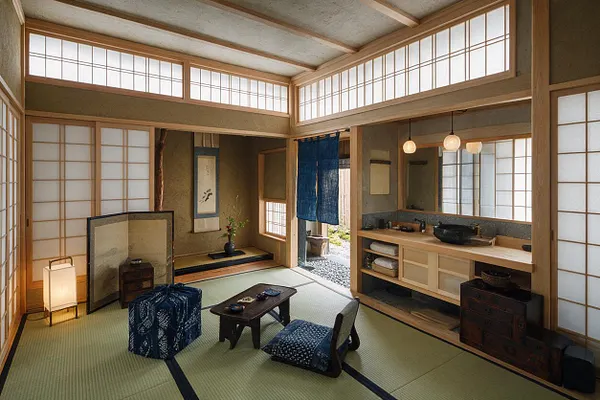 A spacious Japanese Zen-style master bathroom with tatami flooring, shoji screens, light wood framing, and a built-in vanity with a black stone sink. The room includes a low tea table, floor cushions, soft lantern-style lighting, and a blue noren curtain near the bathing area, creating a calm, traditional atmosphere.