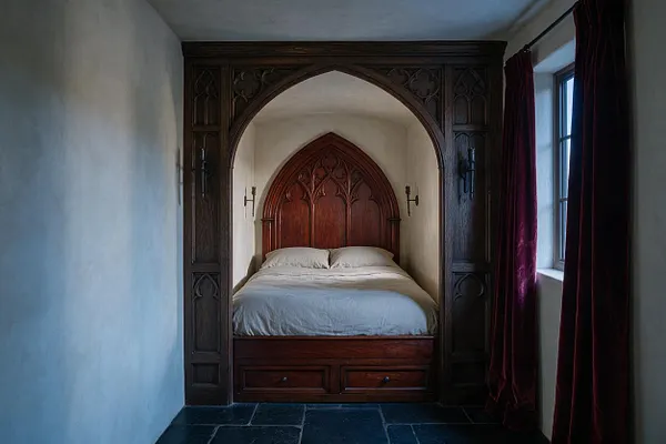 A compact studio apartment bedroom framed by a dark carved wooden Gothic arch and paneling. Inside the alcove is a bed with a tall pointed headboard in rich wood, white bedding, and stone-look dark floor tiles, with deep burgundy velvet curtains by a window on the right.