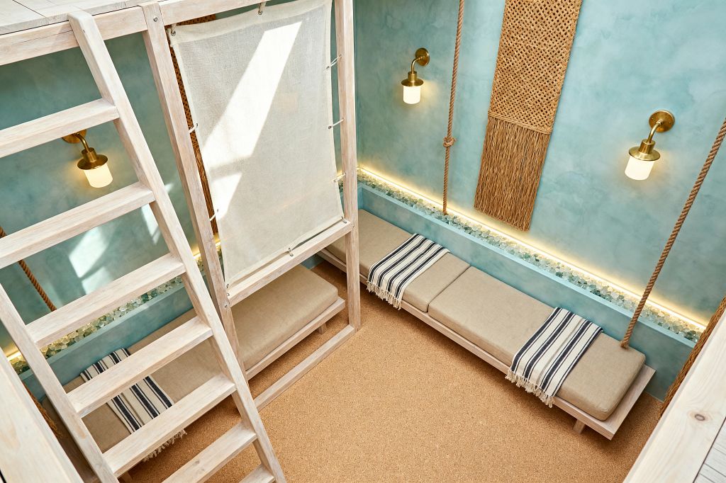 Top-down view of a coastal-style media room with aqua limewash walls and built-in bench seating. Beige linen cushions on a whitewashed wood platform are accented by navy-and-cream striped throws. A trough of sea glass runs along the wall and is lit by warm LED strips beneath brass sconces with frosted shades. Natural rope details and woven seagrass panels add texture, while a cork floor anchors the light, beachy palette.