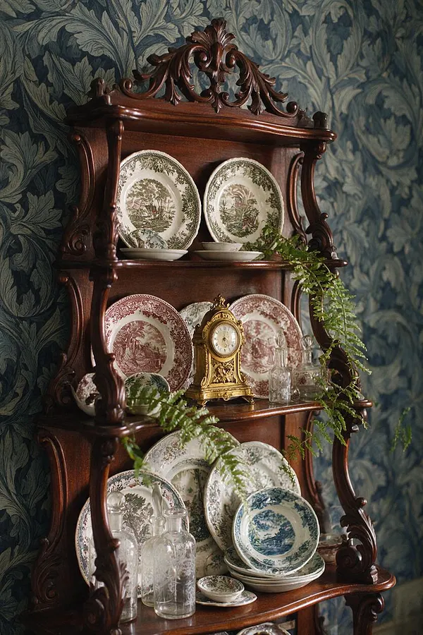 A dark carved wooden Victorian wall shelf holds patterned ceramic plates, a gilded mantel clock, and clear glass bottles. The shelf is set against blue-green floral wallpaper, with trailing greenery draped across the shelves.