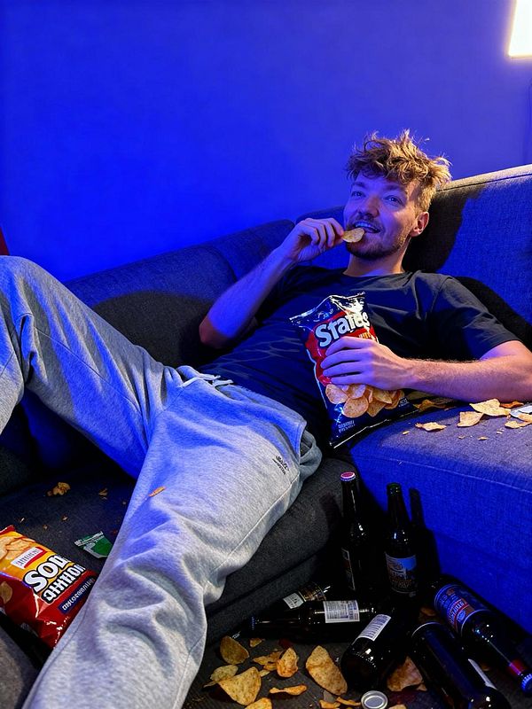 A relaxed individual enjoys snacks while lounging on a couch surrounded by scattered chips and empty bottles.