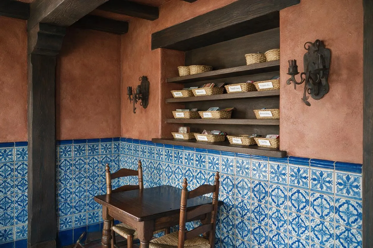 A small Mediterranean-style game room features terracotta stucco walls, dark exposed ceiling beams, and a band of blue-and-white patterned tile wrapping the lower walls. A dark wood table with two woven-seat chairs sits in the corner, while open shelves hold wicker baskets and small labeled items beneath wrought iron wall sconces.