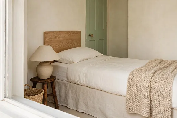 A cozy organic modern bedroom with a single bed dressed in white bedding and a textured beige throw. The room features a natural wood headboard, a sculptural ceramic table lamp on a dark wooden stool, a woven basket on the floor, and a muted sage green door against soft cream walls.