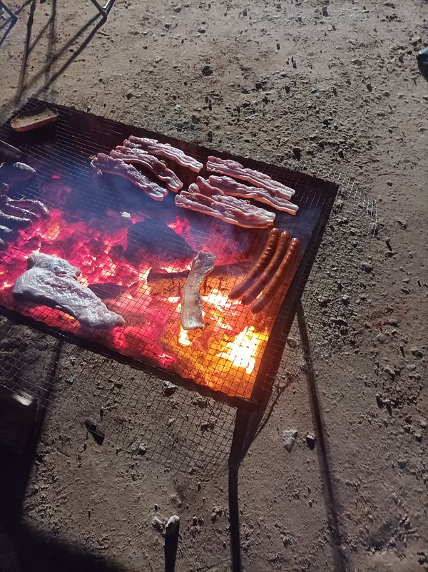 A grill filled with various meats is cooking over glowing coals on a beach.