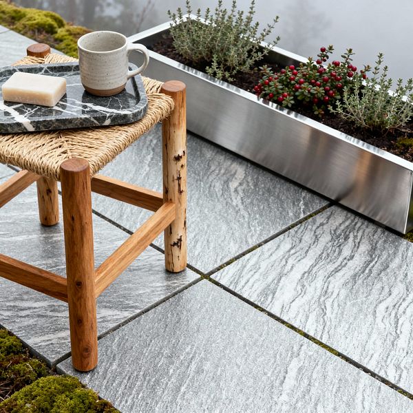 A Scandinavian patio vignette features large gray granite pavers with linear veining and moss between joints. In the foreground is a natural wood stool with a woven seat, topped by a black marble tray holding a speckled gray stoneware mug and a soap bar; along the edge, a brushed stainless-steel trough planter grows thyme and a small shrub with red berries against a soft, misty backdrop.