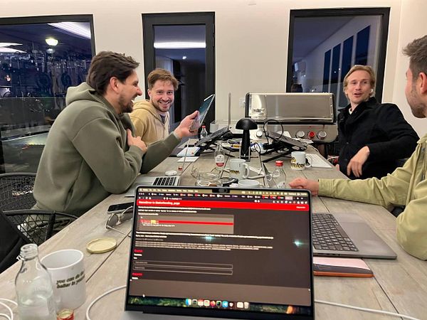 A group of four men are engaged in a lively discussion around a table filled with laptops and drinks.
