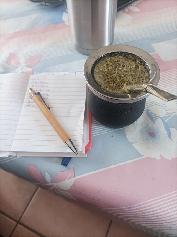 A workspace setup featuring mate tea, a notebook, and a pen on a patterned tablecloth.