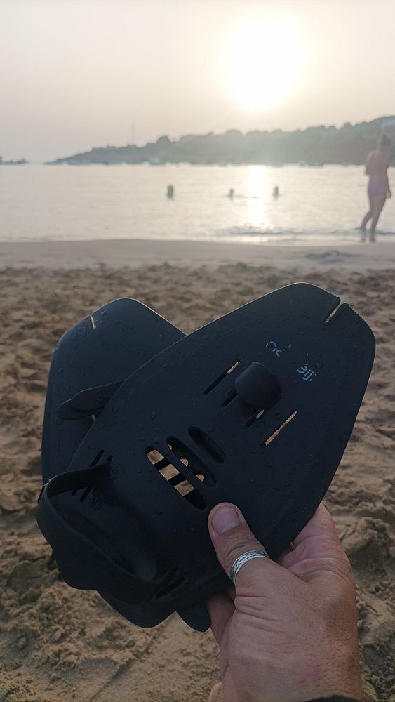 A person holds a pair of black swim fins against a beach backdrop at sunset.