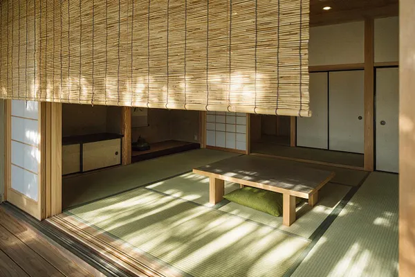 A traditional Japanese tatami room with low wooden table, sliding shoji and fusuma doors, and a woven bamboo blind hanging across the top of the room. Sunlight casts soft striped shadows over the green tatami mats, creating a calm, minimalist Zen atmosphere.