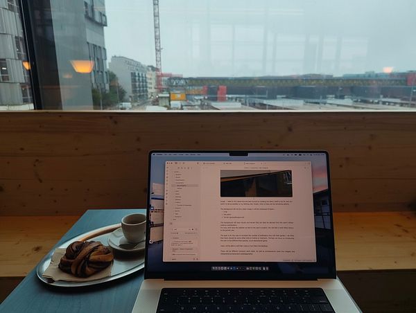 A cozy workspace featuring a laptop, a coffee, and a pastry with a view of a cityscape in Basel.
