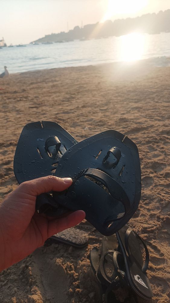 A person holds a pair of swim fins on a sandy beach during sunset.