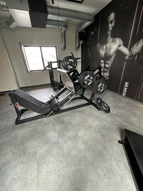 A well-equipped gym space featuring a leg press machine and a wall mural of a muscular man.