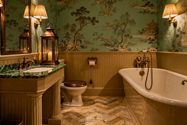 A warmly lit traditional bathroom features seafoam chinoiserie wallpaper above beige beadboard, a white freestanding slipper tub with brass telephone-style faucet, and a cream vanity with a deep green marble countertop and brass taps. A herringbone marble floor, pleated wall sconces, a large gilt mirror, a candle lantern, and a toilet with a wooden seat complete the classic, old-world look.