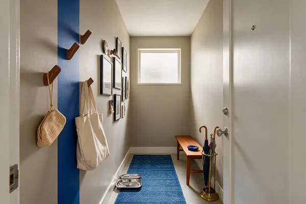 A narrow mudroom with beige walls, a bold vertical blue accent stripe, and a row of wooden wall hooks holding a woven bag and canvas tote. Framed art decorates the left wall, while a small wooden bench, umbrella stand, blue runner, and pet bowls add function near a frosted window.