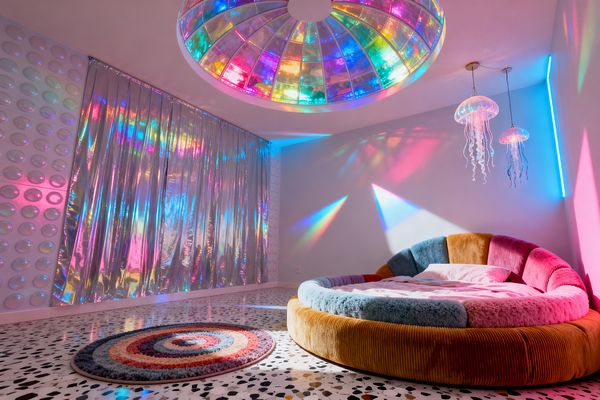 Kids’ bedroom featuring a circular, multicolored upholstered bed on black-and-white terrazzo flooring. A large rainbow-tinted acrylic dome in the ceiling projects colored beams across white walls. Two jellyfish-shaped pendant lights and a slim neon strip glow in the corner. One wall has iridescent foil curtains and another shows raised bubble panels. A round spiral shag rug sits nearby, creating a playful, psychedelic atmosphere in saturated pink, blue, and cyan light.