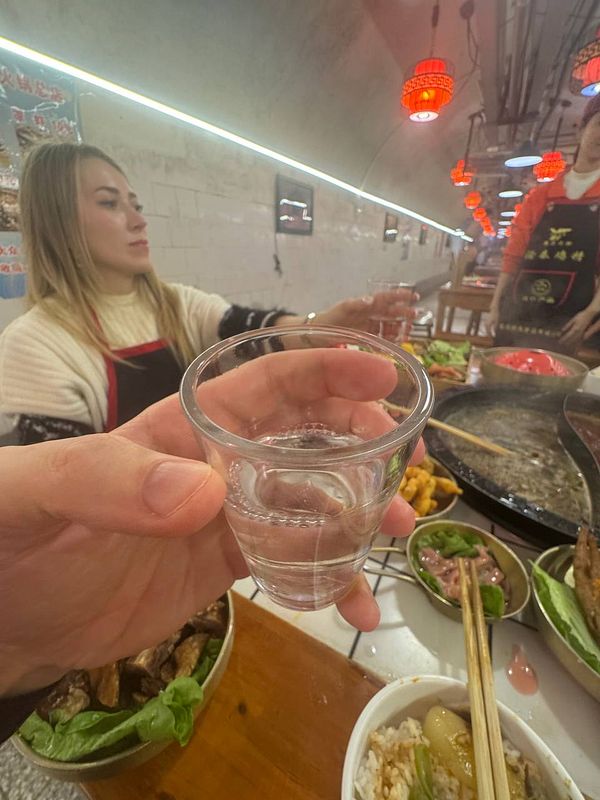 A hand holds a small glass of clear baijiu in a lively dining setting.