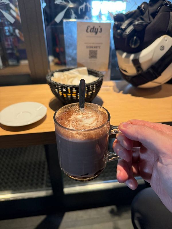A hand holds a glass of hot chocolate in a cozy café setting.
