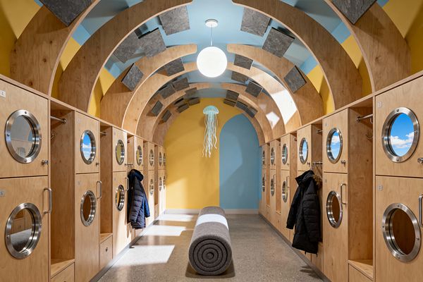Long, tunnel-like mudroom with curved plywood arches overhead and walls split between sunny yellow and sky blue. Light wood lockers line both sides with round stainless porthole doors showing cloud imagery, plus hooks and handles; a rolled gray felt bench rests on polished concrete below a glowing globe and a jellyfish-like pendant, while rectangular gray acoustic panels appear to float across the ceiling, creating a playful, surreal atmosphere.