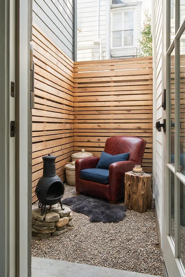 A small rustic backyard seating nook enclosed by horizontal wood slat fencing, with a burgundy leather armchair, blue cushion, black chiminea, woven baskets, and a tree stump side table. The ground is covered in gravel with a dark sheepskin-style rug, creating a warm, intimate outdoor retreat.