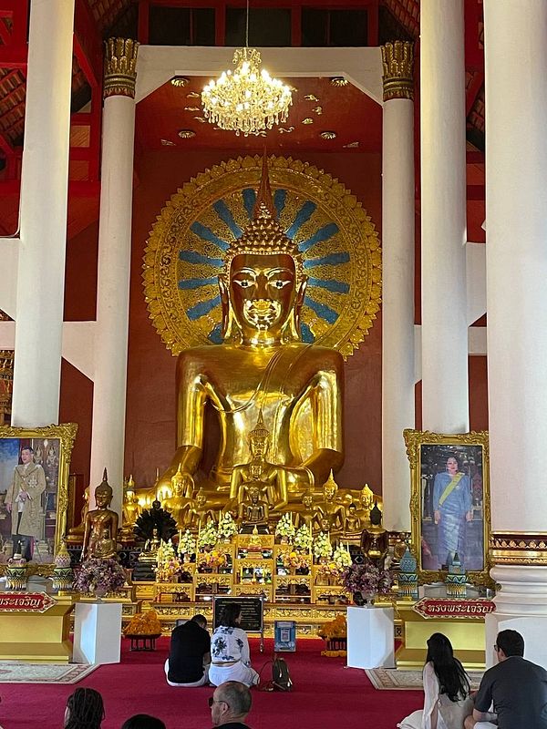 A serene interior of a temple featuring a large golden Buddha statue and worshippers in prayer.
