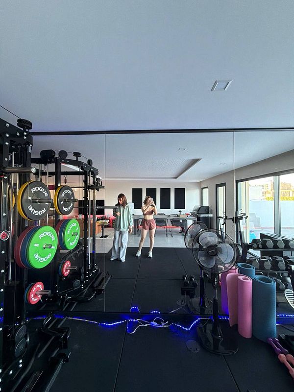 A woman wearing a white sports bra and black leggings exercises barefoot, lifting a large blue kettlebell on a wooden floor in a light-filled room with a window and green plants.