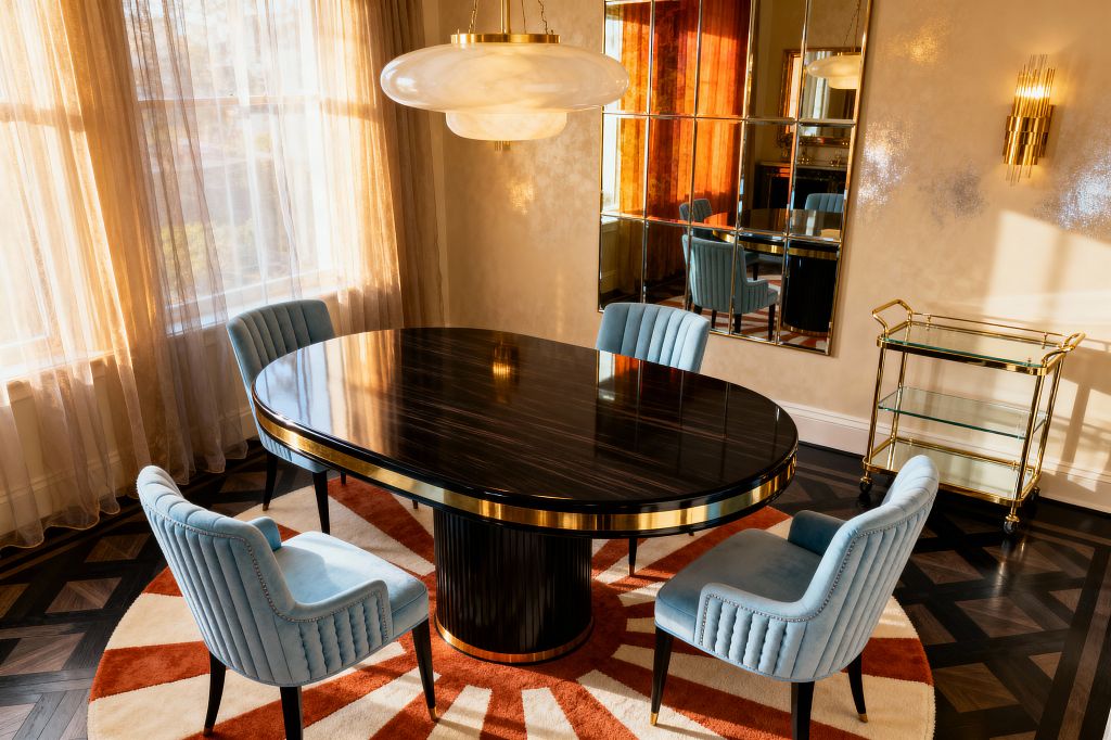 A vintage glam dining room features a glossy oval ebony table with brass trim and a fluted pedestal, surrounded by four powder-blue, channel-tufted velvet chairs on a round sunburst rug of burnt orange and cream over dark parquet flooring. Champagne walls host an antiqued grid mirror, a brass sconce, and a glass-and-brass pendant, while sheer golden drapes diffuse daylight and a brass-framed glass bar cart stands to the side.