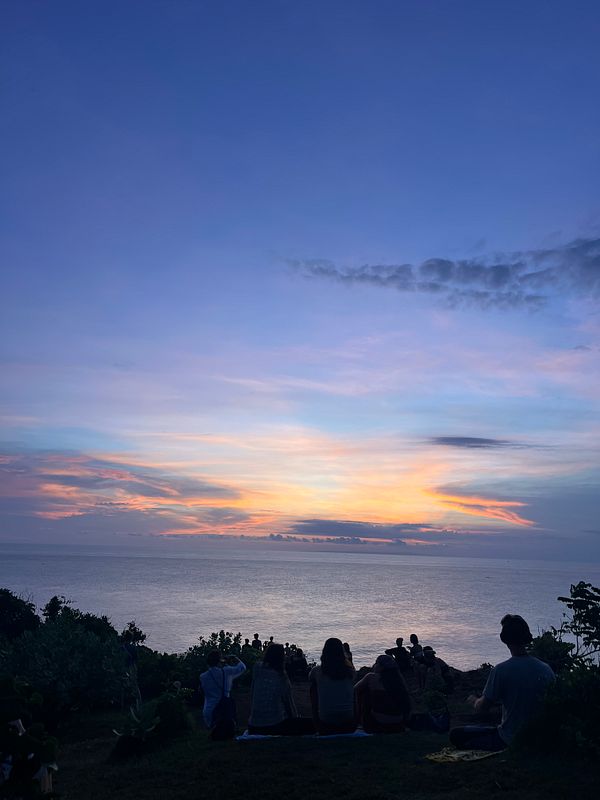 A group of people sits on a grassy area overlooking a serene ocean sunset in Uluwatu.