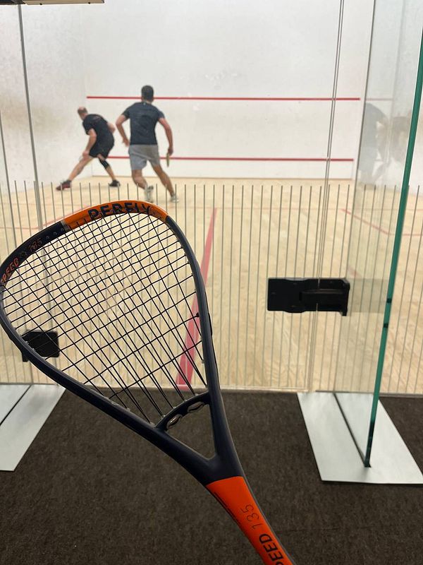A squash racket is prominently displayed in the foreground while two players engage in a match in the background.