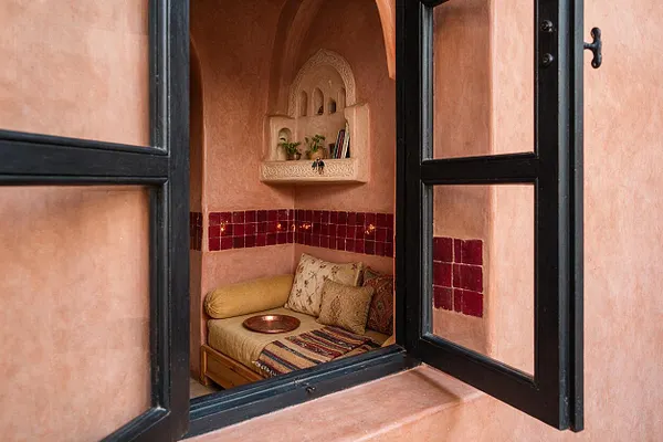 A small Moroccan-style sunroom nook is framed by open black metal windows and soft pink plaster walls. Inside, a built-in daybed with patterned cushions, a striped textile, red tile accents, and an arched wall shelf create a warm, intimate seating area.
