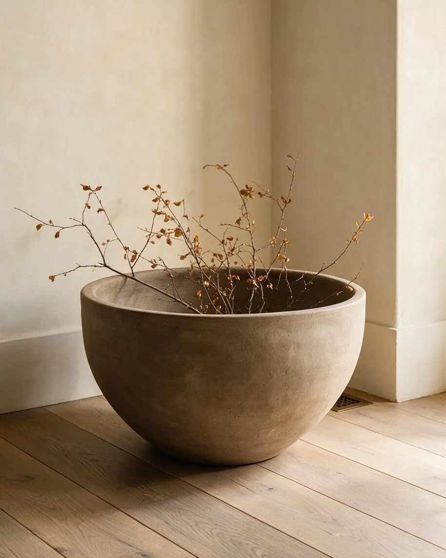 A large rounded planter or sculptural bowl sits on a light wood floor in a quiet corner. It holds delicate bare branches with small orange-brown leaves, set against soft cream walls and natural daylight.