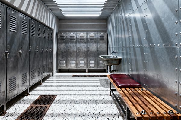 An industrial mudroom features rows of galvanized metal lockers on the left and back walls, a riveted sheet-steel wall on the right, and a wall-mounted cast-iron utility sink. A slatted skylight casts bold shadow lines across a speckled terrazzo floor with inset floor grates, while a slatted wood bench with a burgundy cushion provides seating, creating a durable, easy-to-clean space with a cool gray palette warmed by natural wood.