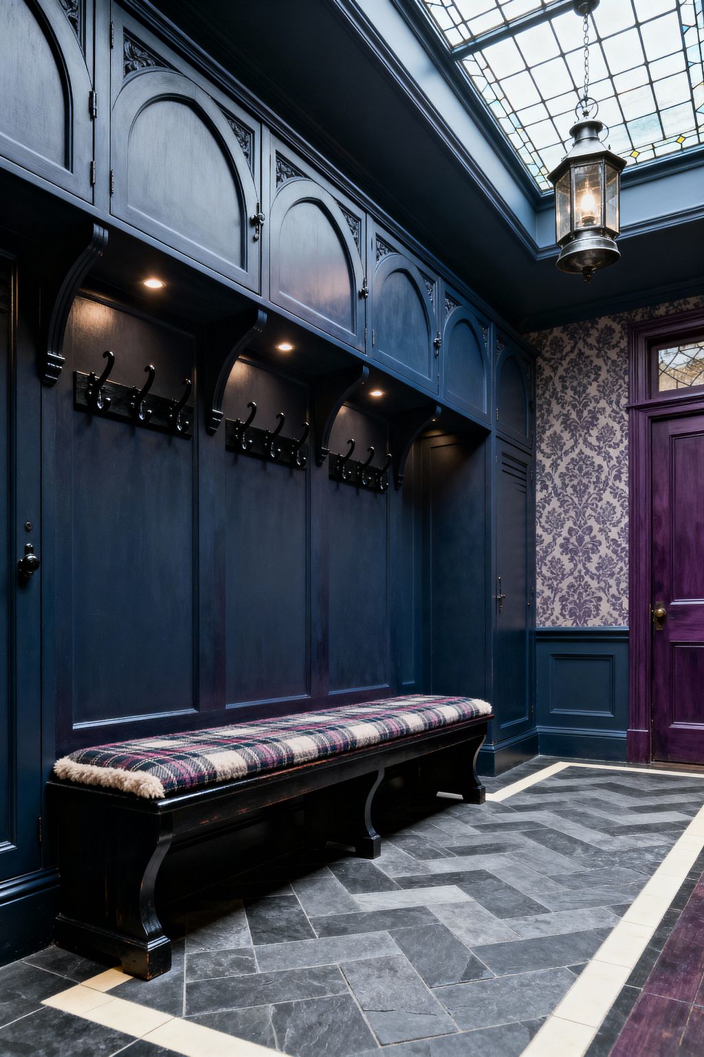 A richly detailed mudroom features deep navy built-in lockers with arched cabinet doors, carved brackets, and rows of black iron hooks. A long bench with a plaid wool cushion sits above a dark gray stone herringbone floor edged with a light border. To the right, purple damask wallpaper rises above matching wainscoting and an aubergine door. Overhead, a large leaded-glass skylight and a hanging lantern cast light onto the cabinetry and integrated downlights.