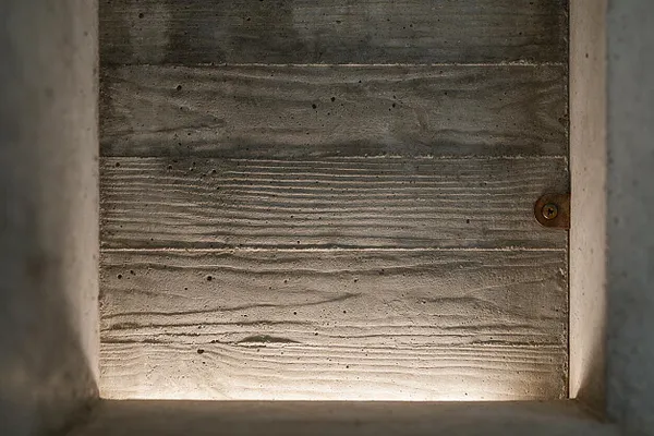 A close-up view of a recessed concrete panel with horizontal wood-grain impressions and small air holes, framed by rough concrete edges. A single exposed fastener appears on the right side, emphasizing the raw, unfinished Brutalist character.