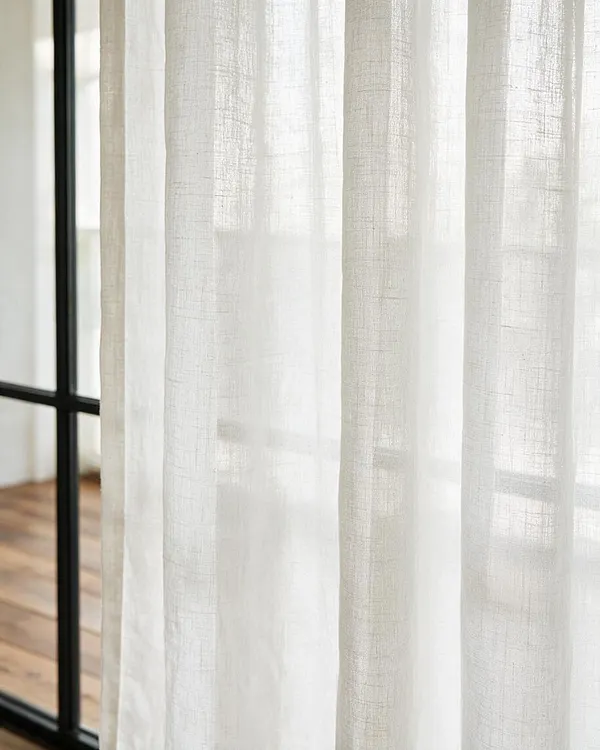 A close-up of sheer white linen curtains hanging beside a black metal-framed window. Soft daylight filters through the lightweight fabric, with warm wood flooring faintly visible in the background.